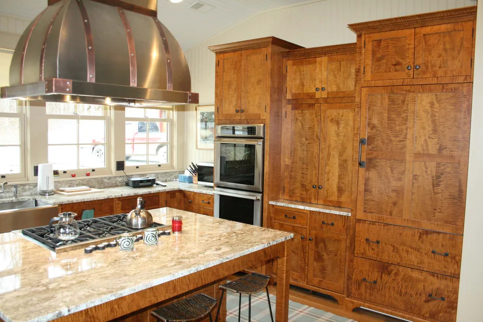 Kitchen with highly figured wood cabinetry, copper-colored straps on a domed hood, a paneled refrigerator, and dark countertops — the Boat House project.
