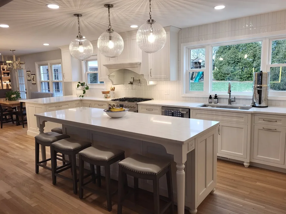 White furniture-style kitchen island with turned legs and saddle-seat stools under ribbed glass globe pendants.